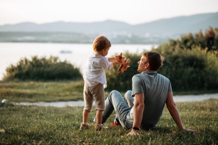 Vater sitzt mit seinem Sohn im Gras am See und spielt mit einem Holzflugzeug