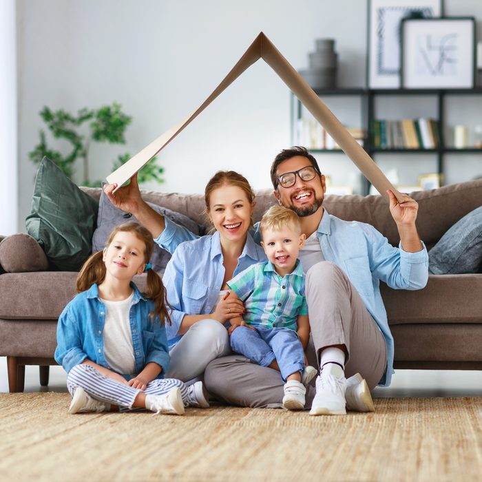 Vierköpfige Familie sitzt lachend zusammen und hält ein angedeutetes Hausdach schützend über sich